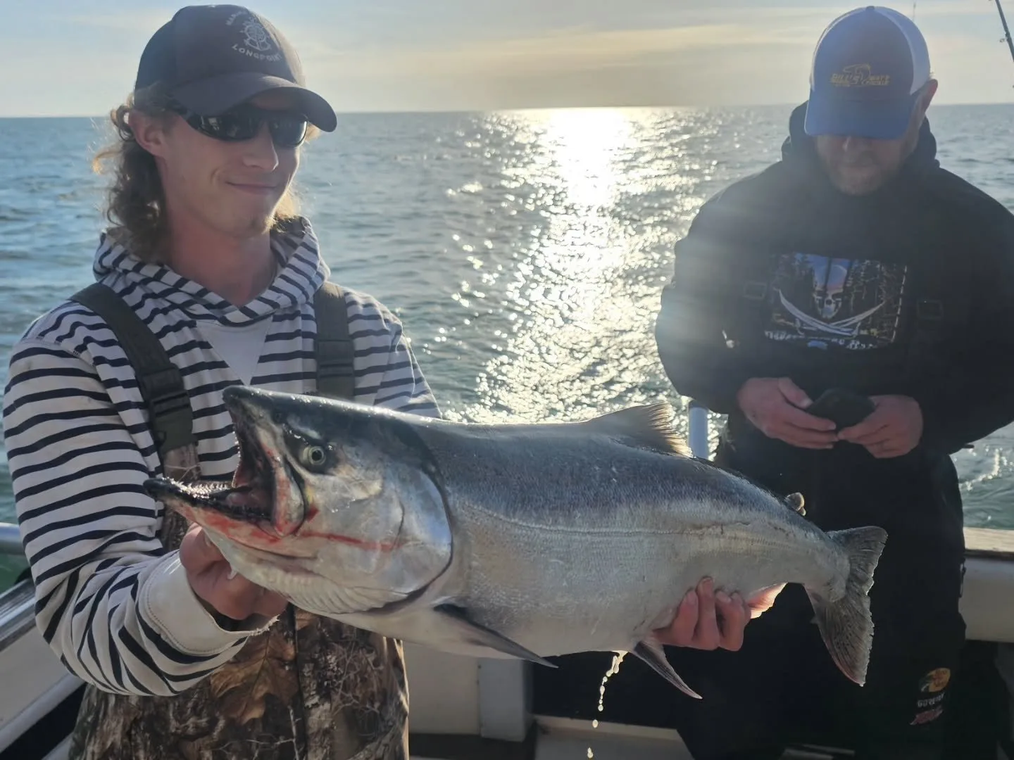 Two anglers aboard a King Slayer boat, one holding a chinook salmon, backlit by the sunset